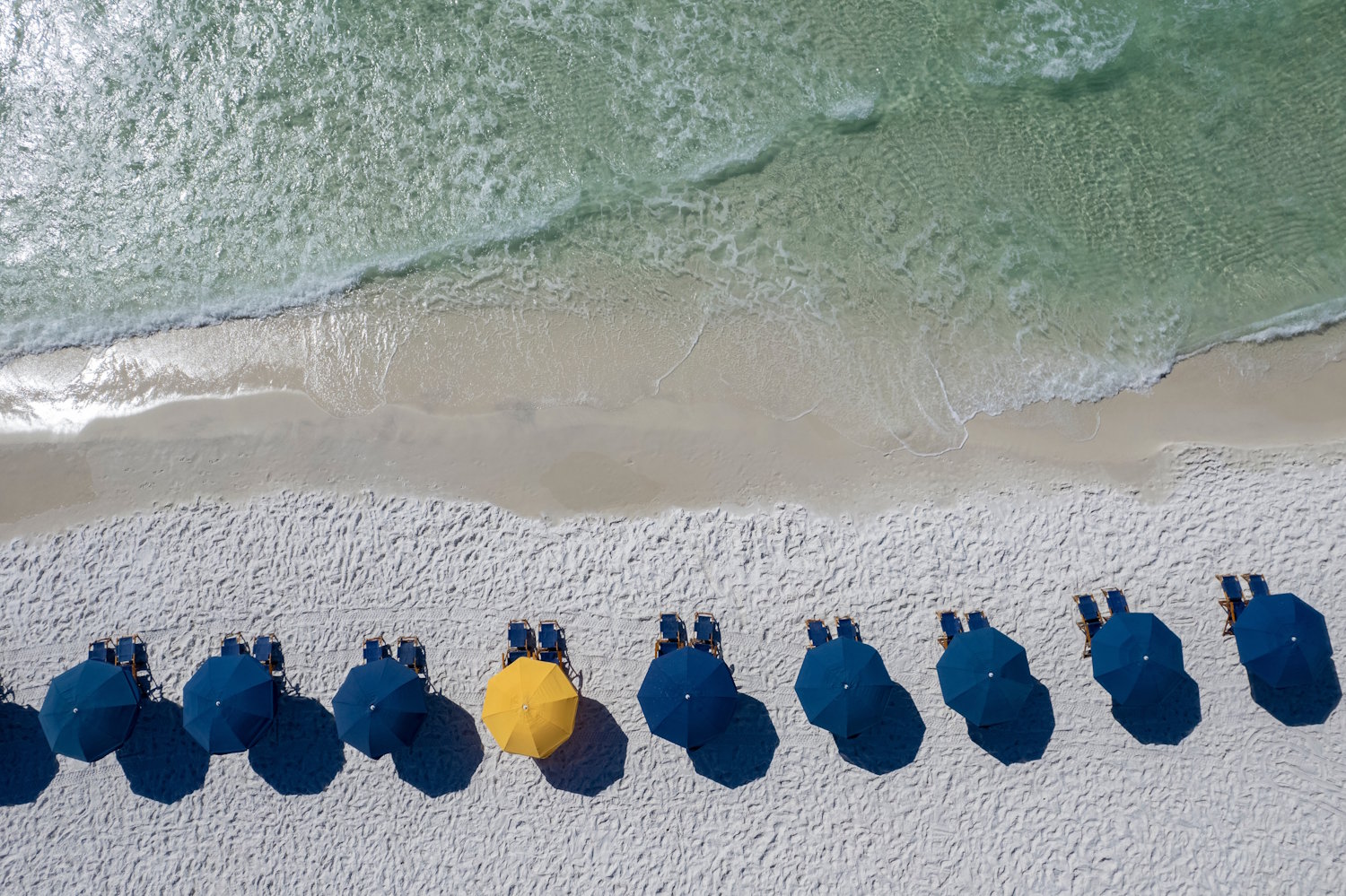 Aerial view of a row of blue beach umbrellas on white sand along the emerald Gulf water in the Florida Panhandle